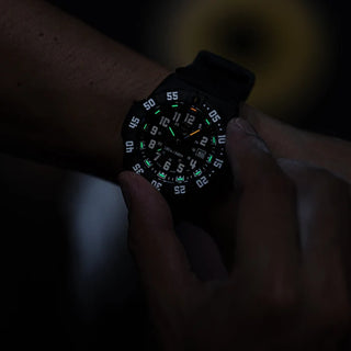 A close-up of a black tactical watch with a glowing green and white luminescent dial, worn on a wrist in a very dark environment.