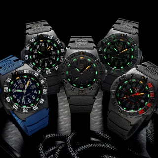 Five tactical-style wristwatches arranged together on a dark surface against a black background. Four watches have black or dark grey cases and dials with glowing green accents, and one watch on the left has a bright blue strap with a dark dial and green/orange accents.