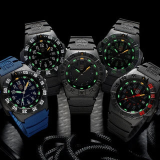 Five tactical-style wristwatches arranged together on a dark surface against a black background. Four watches have black or dark grey cases and dials with glowing green accents, and one watch on the left has a bright blue strap with a dark dial and green/orange accents.